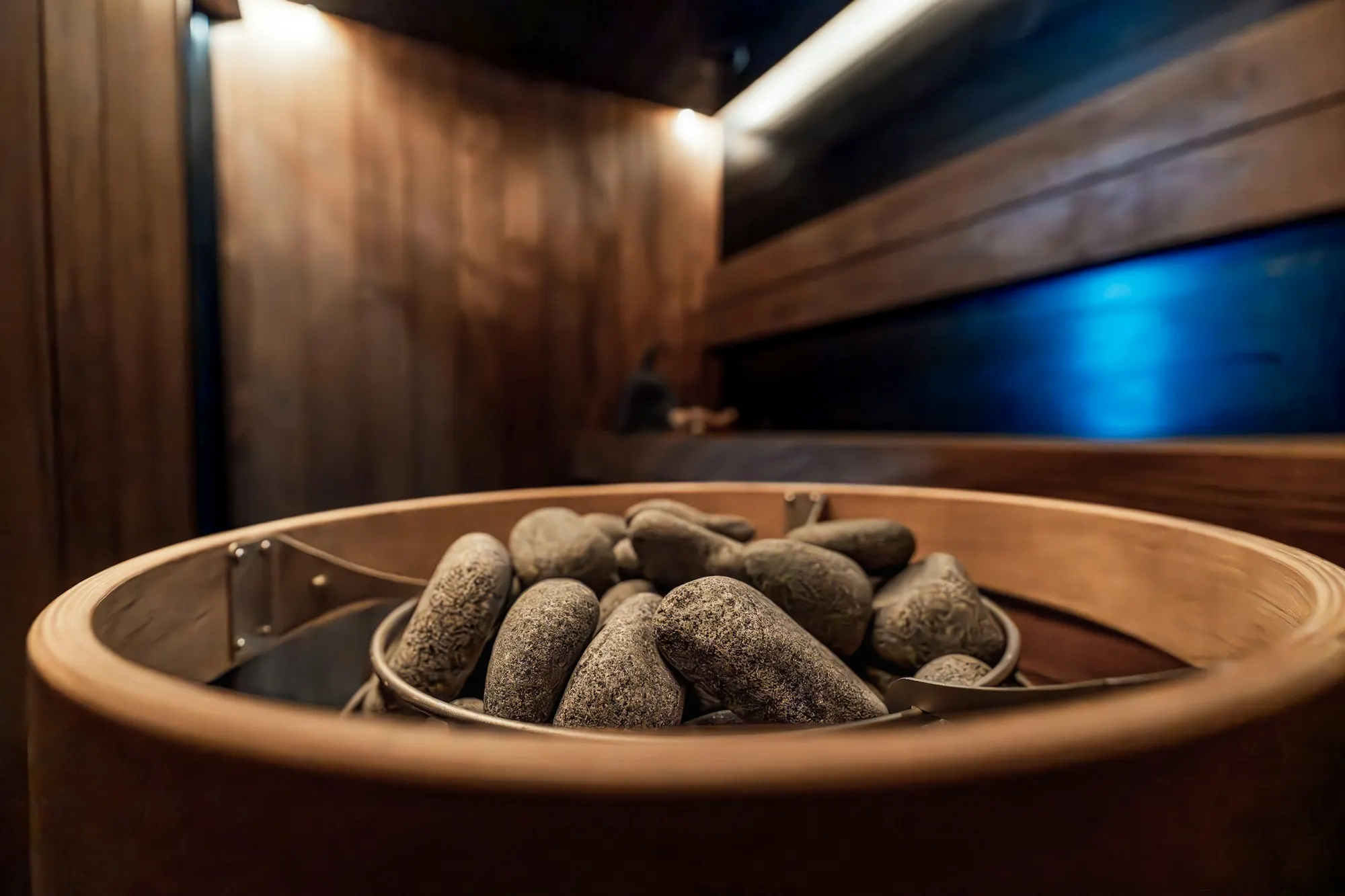 Close view of loose sauna stones resting in the steel basket of a round sauna heater, lit warmly against dark timber.