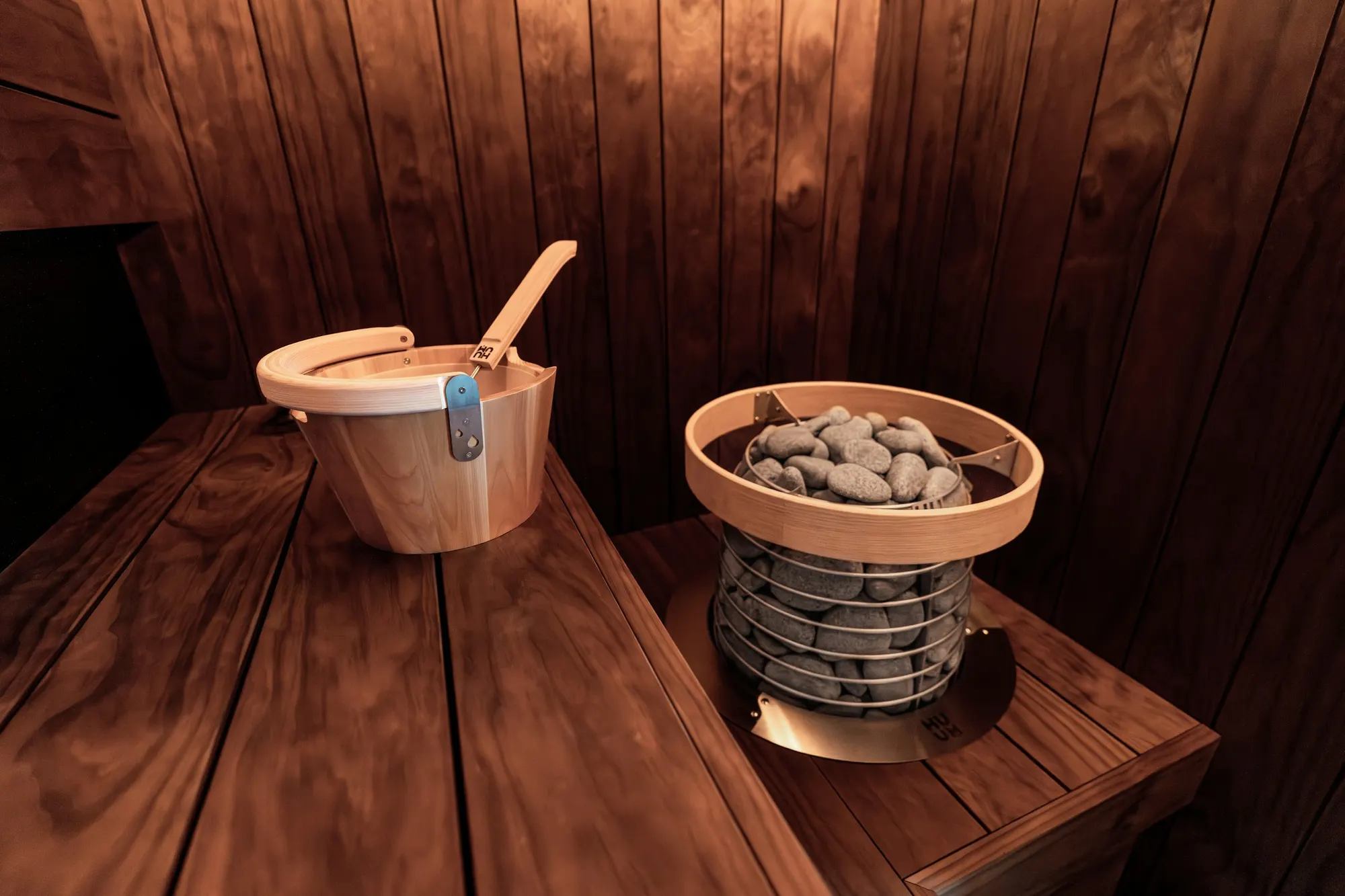A dark timber sauna interior: a wooden bucket and ladle on the bench beside a round heater filled with sauna stones.